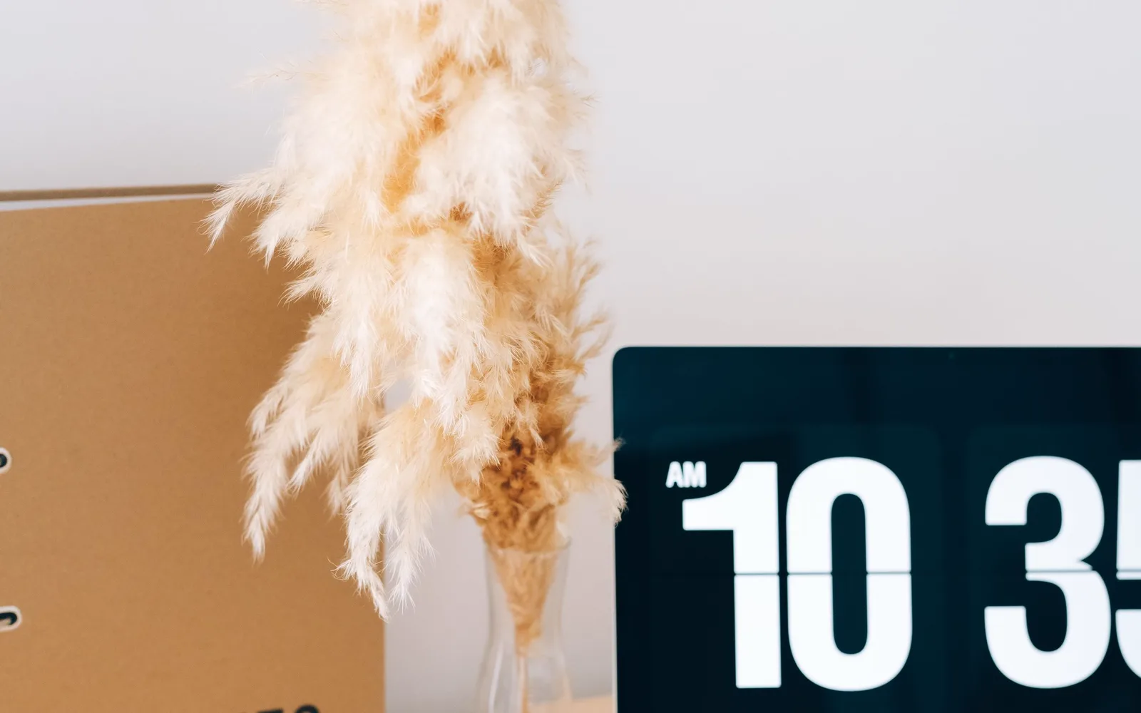 A desk still life with a flip clock, a binder and dried grass in a vase.